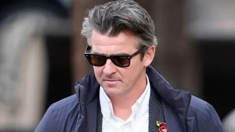 PA Media Joey Barton seen walking into court. He has medium-length black and grey hair and wears sunglasses. He also wears a blue coat over his suit, with a red poppy badge.
