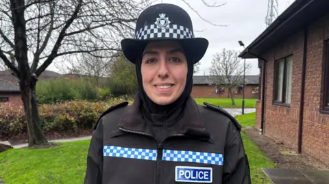 PC Seher Nas in black police uniform including a hijab and police hat, stood in front of a single-storey brick building and a grass lawn. 
