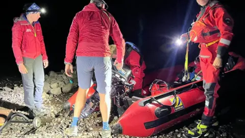 Keswick Mountain Rescue Team members, all wearing red emergency clothing, prepare to send off a rib boat into Thirlmere. There are two rescuers wearing red diving suits and it is pitch black and late at night. 