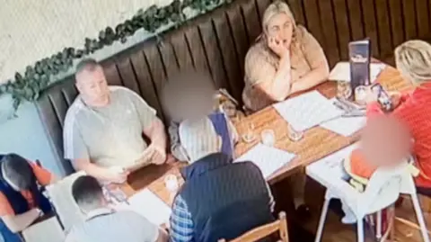 The family at Bella Ciao sitting around a wooden table. Bernard and Ann are sitting on a long bench with a leather brown back, while others sit opposite them, including a child in a white high chair. 