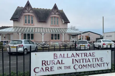 A large detached light pink house with a dark grey roof. Cars are parked in front of it and a black iron fence is in the foreground with a sign which says 