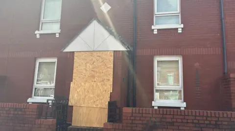 The front door of a red bick house has been boarded up. All of the blinds in the front four windows are pulled. 