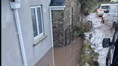 Jennie Spicer The photo shows a flooded cottage with brown water pooled against the exterior walls, reaching several inches deep. The water has risen up to the doorstep and is spreading along a narrow path between the house and a stone wall. The stone wall beside the property is also wet, with water flowing along the adjacent lane where several vehicles are parked or attempting to pass. The road itself appears heavily flooded, with water streaming past the cottages and creating a fast, shallow flow. 