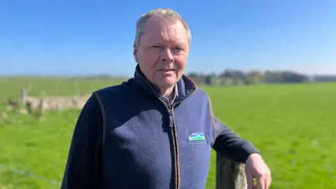 BBC news NFU Scotland president Andrew Connon, with short grey hair, wearing a blue jumper and blue gilet leaning against a wooden post with a large field in the background