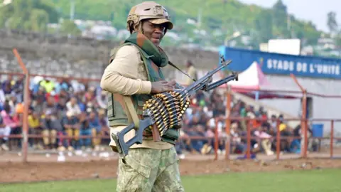 Göktay Koraltan / BBC A M23 fighter holding a gun with lots of bullets wrapped around it. He is standing in a stadium with people seated behind him - Goma, DR Congo