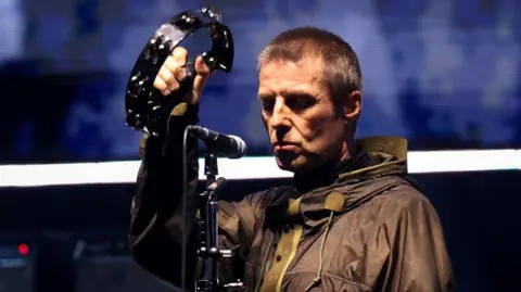 Reuters Liam Gallagher, wearing a raincoat, has a sombre expression as he holds up a tambourine during an Oasis reunion concert