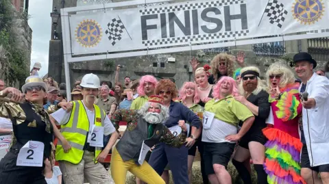 Georgie Faulkner-Bryant About 13 people wearing fancy dress strike silly poses beneath the finish sign at the top of Gold Hill. Among them is a man wearing a multicoloured flamenco dress and a long blond wig, a person in a King Arthur face mask with a beard and flowing grey hair and a woman in a gold top and top hat.