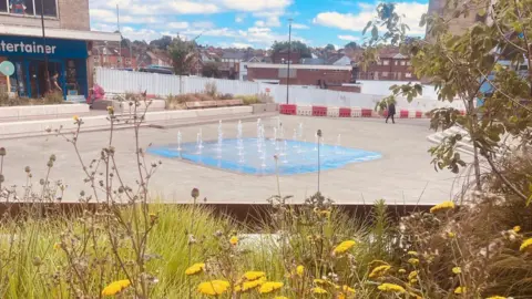 Somerset Council Water jets spout upwards from a patch of blue flooring among paving stones in an open space. Behind is half of a shop, and many red-brick houses. In the foreground there are yellow flowers and green plants.