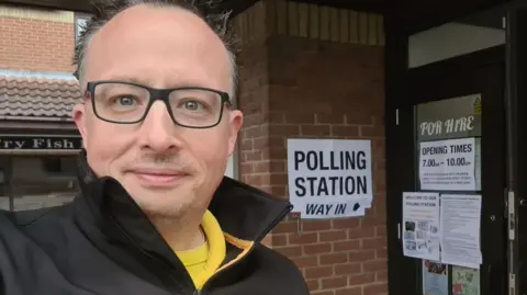 Andy McGuinness Andy McGuinness with short dark hair, taking a selfie while wearing a black jacket and yellow T-shirt. He is standing in front of a brick-built hall which is displaying a "polling station - way in" notices. There are further notices attached to the black doors.