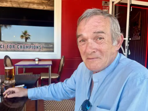 A man sitting outside a bar holding a cigarette, with a pint of lager on the table. He is wearing a light blue shirt with a grandad collar and has a pair of blue-tinted sunglasses tucked into his shirt buttons.