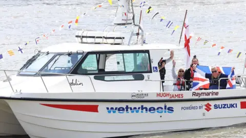 Lia Toby/WetWheels A boat decorated with flags and bunting, with Mr Holt holding up a flag on the back of it