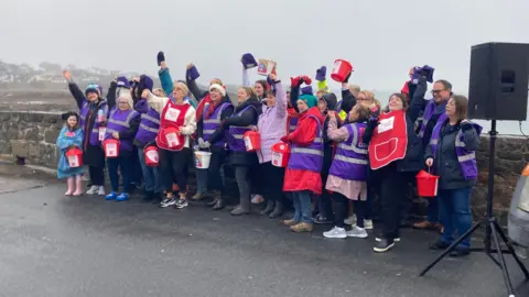 A group of people wearing purple vests hold red fundraising buckets up as the stand in front of a sea wall.