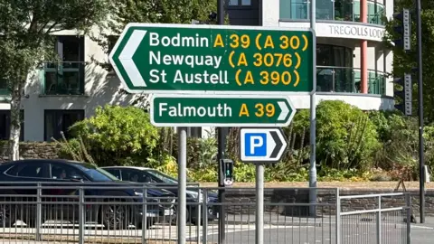 Two green arrow road signs on top of one another - one points to the left directing drivers towards Bodmin, Newquay, and St Austell and the other points to the right towards Falmouth. 