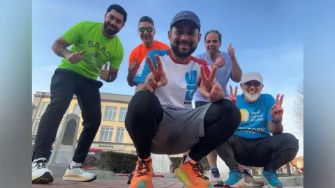 Ramadan Runners Five men posing for a photo in their bright running gear. Two wearing baseball caps crouch on the floor and hold up their fingers in a peace sign, while the other three lean forwards with their thumbs up. They are in a town centre and the sky is blue behind them. 