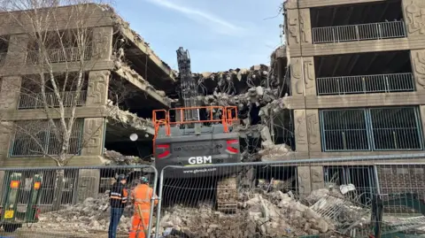 Grey concrete car park being demolished. There is an orange bulldozer in the centre of the picture which is tearing down the car park. There are piles of rubble on the ground. Two men are standing looking at the car park with their back to the camera, one is wearing an orange boiler suit and a hard hat, the other man is wearing jeans, a blue top and a black hard hat