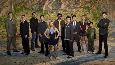 Getty Images Panettiere, with straight long blonde hair and a neutral expression, wearing a blue and navy cheerleader's kit, is seen stood in the centre of the season 2 cast of Heroes in a promotional picture for the show.