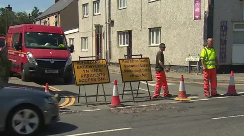 BBC Road in the centre of Newborough with yellow closure signs, cones and traffic management staff