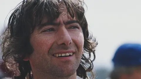 Getty Images Joey Dunlop has long dark brown hair with a fringe, he has dark eyebrows and facial stubble. He is wearing a white racing suit and is looking to his left, the background is blurred.