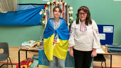 Shariqua Ahmed / BBC Iolia and Tesilisa smiling for the camera in front of the Ukraine stall with Lolia holding the country's flag around her