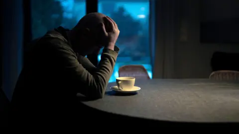 A man is sitting alone at a kitchen table in a dim room. There is a coffee cup in front of him. He has his head in his hands and looks down. It's dark outside, as it can be seen through the windows