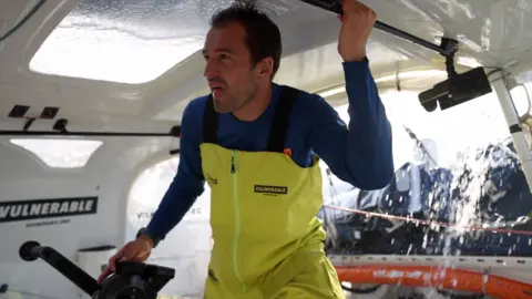 Mark Lloyd/TR Racing A man in a yellow sailing trousers and a blue top in a cockpit of a yacht with water pouring off the top of the cabin behind.