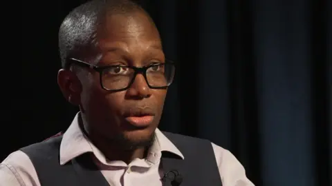 A black man with closely cropped hair, a short goatee and black-rimmed glasses is mid-speech, talking to someone out of frame. He wears a white shirt and navy waistcoat with a small lapel microphone clipped to it. He is sat against a faintly lit black curtain.