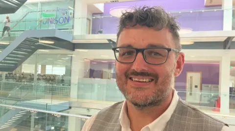 Vikki Irwin/BBC A man inside a glass building. In the background is the central atrium with flights of stairs. He is wearing a checked waistcoat and a white shirt. He is smiling at the camera.