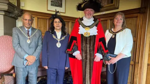 Four people standing side by side in a council chamber. The centre figure, a man, is wearing red ceremonial robes and a large gold chain with an ornate medallion. A man on the left is in a grey suit with a light shirt and tie, and is also wearing a ceremonial chain. Second from left a woman is wearing a dark, tailored suit and a ceremonial chain. On the far right a woman is dressed in a dark outfit with a light-coloured jacket and is carrying a small clutch bag. A medallion on a chain is also worn.