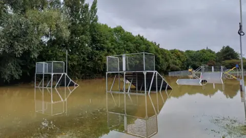 Metal ramps of a skate park partially under water, with trees and a playground in the background.