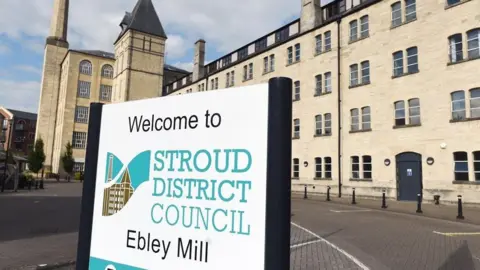 A photo taken outside of the offices of Stroud District Council with Cotswold stone buildings in the background and a sign.
