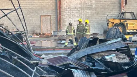 Fire fighters stand in a scrap metal yard with large sheets of metal lying around in piles. 