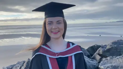 Francesca Murphy Francesca Murphy wearing graduation apparel  and cap, standing, looking into camera with Swansea formation  down  her. She is smiling.