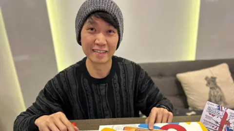 A man in a grey and black patterned jumper, wearing a grey beanie hat. He is smiling and has his hands on a table, where board games can just be seen. A sofa with a cushion featuring a print of a dog is just behind him.