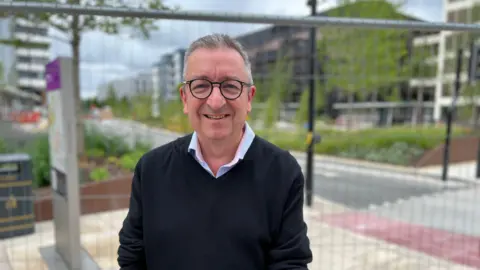 A bespectacled man smiles whilst standing in front of a metal fence with a newly constructed bus boulevard and various trees and buildings in the background.