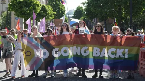 A line of eight people at the front of a protest march are holding a banner with the words 'No going back!' The banner features the colours of the transgender flag and pride flag