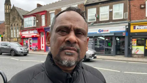 Masaid Ahmed is looking directly at the camera with a neutral expression on his face. His short, dark hair and beard are greying, and he is wearing a puffy black coat. There are colourful shop fronts behind him and a church.