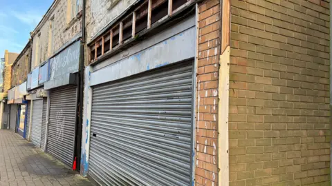 A row of shopping units in Stanley's Front Street with their shutters down. The paint is crumbling off the walls in some places.