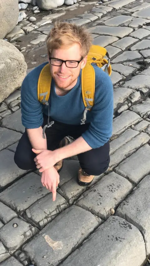 Matthew Myerscough Matthew, wearing dark rimmed glasses and a yellow backpack, standing on rocky ground pointing to a fossil on the stones in front of him. 