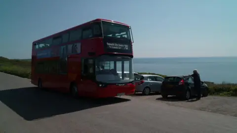A red double-decker bus on a road, with the sea and sky behind. There is a row of parked cars directly behind the bus on the side of the road.