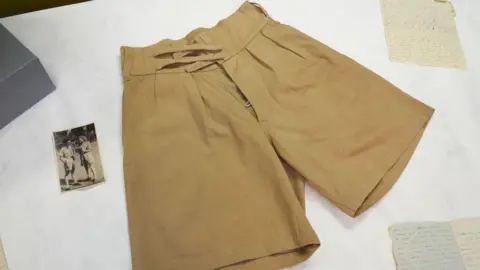 PA Media Khaki shorts worn by Dame Vera Lynn are laid out flat on a table next to a black and white photograph of the singer wearing them as she stands outside and talks to a man. There is a handwritten letter in the corner of the photograph taken in the museum.