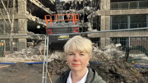 Lara King/BBC Woman with short blonde hair wearing a green coat and silver hoop earrings standing in front of a concrete car park which is being demolished. In the centre of the picture behind her is an orange bulldozer, which is demolishing the grey coloured car park. There are piles of rubble on the ground and a metal fence.