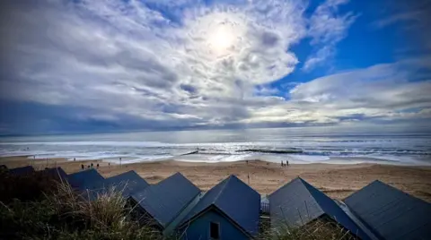 A row of beach huts look out on to a beach. A few people can be seen on the sand in the distance. The sky is a mix of clear blue, white clouds and more threatening grey clouds rolling in from the left