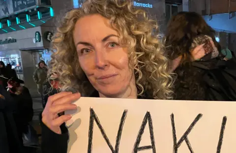 A woman with shoulder-length, blond, curly hair appears in close-up. She is holding a sign although the words cannot be seen in the image. She is standing outside with other people during a protest.