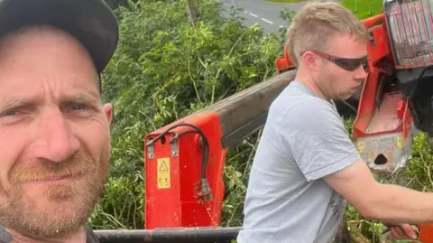 Northumbria Police Daniel Graham is in the forefront of the shot with a ginger beard and wearing a cap. Behind him, Adam Carruthers is standing next to some tree felling equipment. He has fair hair, is in his 30s, wearing sunglasses and a grey top. He's looking at the equipment, not at the camera.