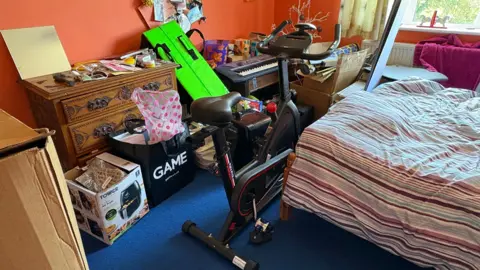 Martin Heath/BBC A bedroom with a bed to the right and boxes and bags full of belongings on the floor to the left. The belongings include an exercise bike and a keyboard. There is a window in the background.