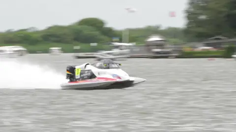 A single white and black powerboat races from left to right with water spraying behind it. In the background are blurry buildings and trees, and flagpoles.