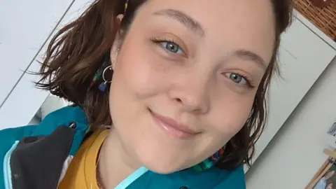 Eleanor Goodman Eleanor Goodman taking a selfie and looking at the camera. She has short brown hair and is wearing earrings. She is smiling and is wearing a yellow top with a blue jacket.