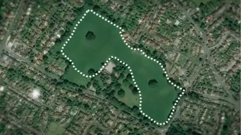 A bird's eye view of the land, shown with a dotted white line around it. There are lots of houses and trees all around the green space. The land itself is vaguely rectangular, and solid green with grass, with a couple of trees in the middle. 