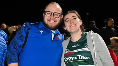 Man with glasses and beard in blue Bath rugby top and woman in Leicester Tigers shirt with grey hoody