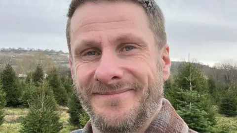 Chris Hurcombe, who has a brown and grey beard, is smiling while standing outside by trees. 